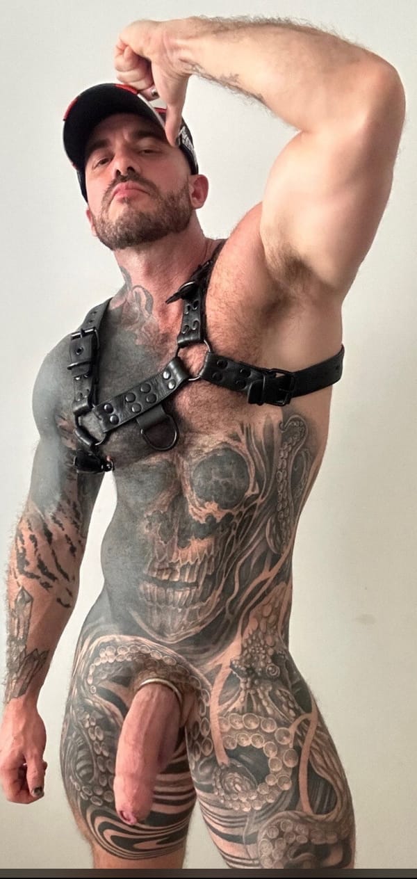 A heavily tattooed man, adorned in a black leather harness and a baseball cap, strikes a powerful pose, showcasing his muscular physique and intricate body art.
